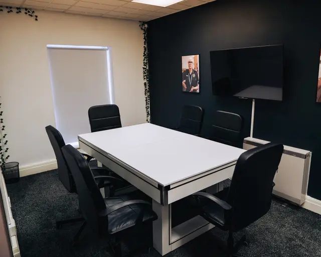 Modern boardroom setup with white table, black chairs, and wall-mounted TV screen.