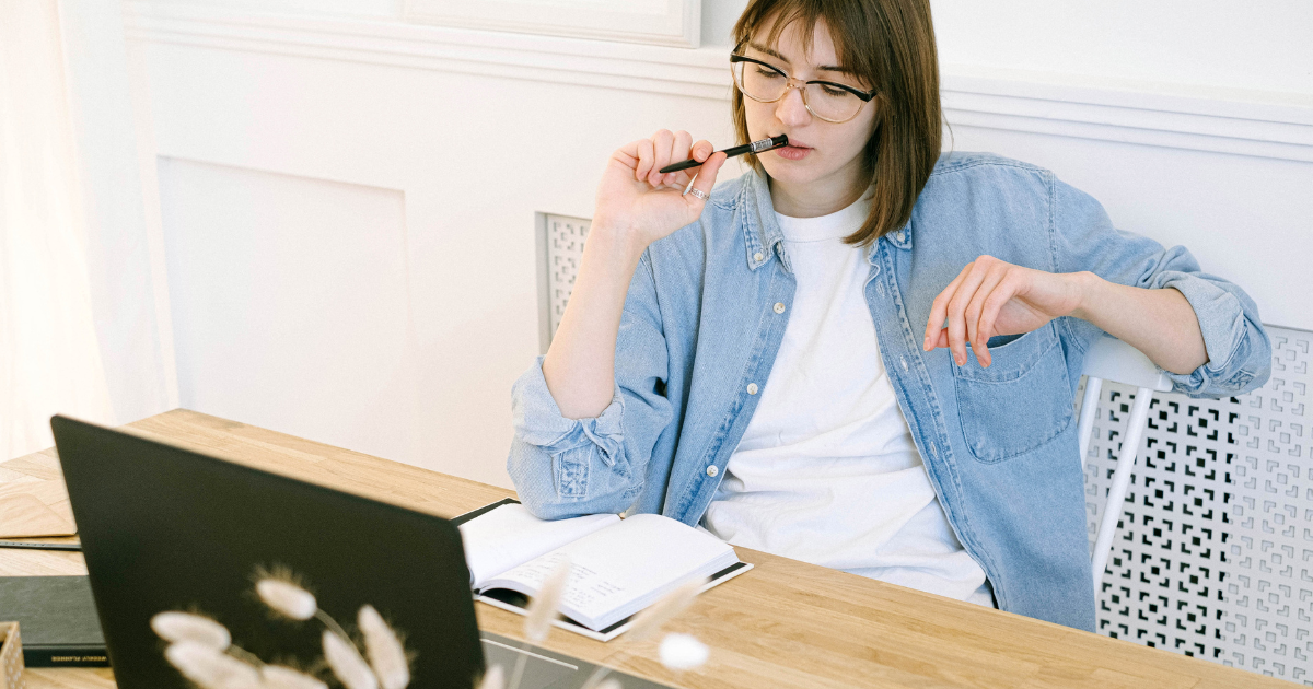 A woman in glasses sitting at a desk, thinking while holding a pen, with a laptop and open notebook in front of her.