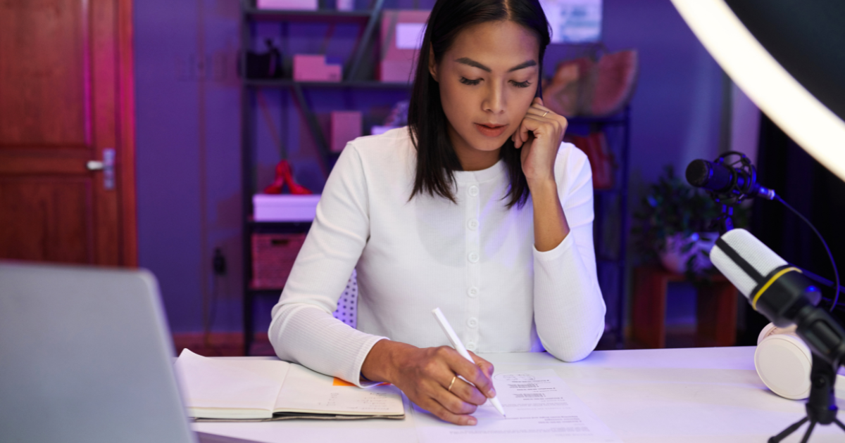 Woman writing on paper at a desk in a studio with microphones, lighting, and creative background setup.